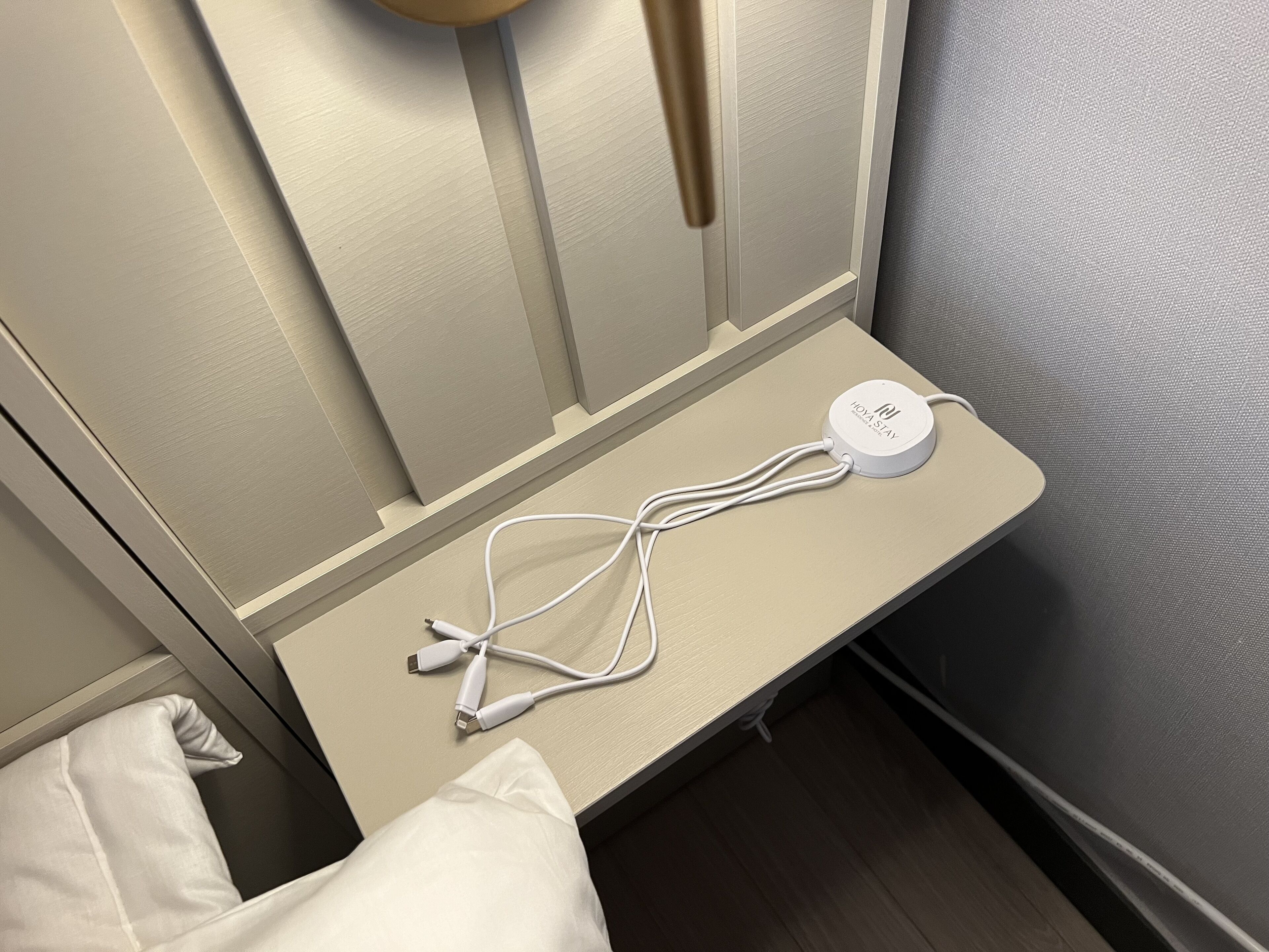 Une table de chevet avec une station de recharge blanche et des câbles, un oreiller et une lampe fixée au mur.