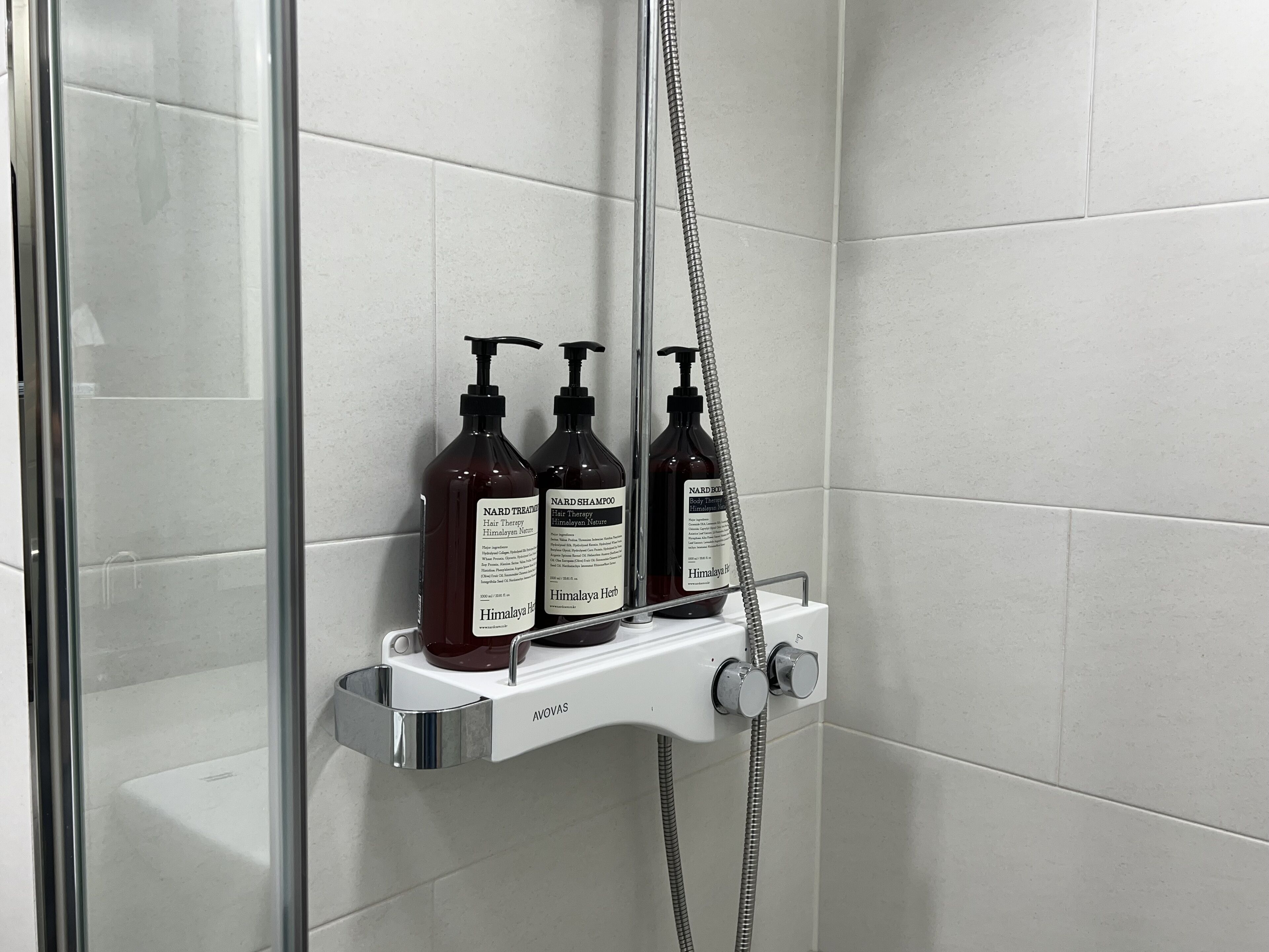 Douchegedeelte met een glazen deur, een witte betegelde muur en twee flesjes Nard Shampoo op een plank.