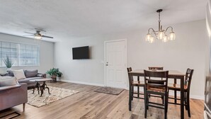 Amplia sala de estar con ventilador de techo, televisor de pantalla plana montado en la pared, comedor con mesa y sillas de madera, y un ventanal con persianas.