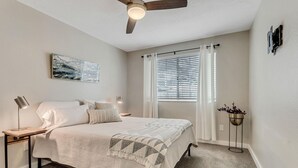Un dormitorio con cama, mesita de noche, lámpara de pie, ventilador de techo, ventana con cortinas e imagen colgada en la pared.