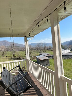 A porch with a swing chair, a view of a rural landscape with mountains, and a barn.