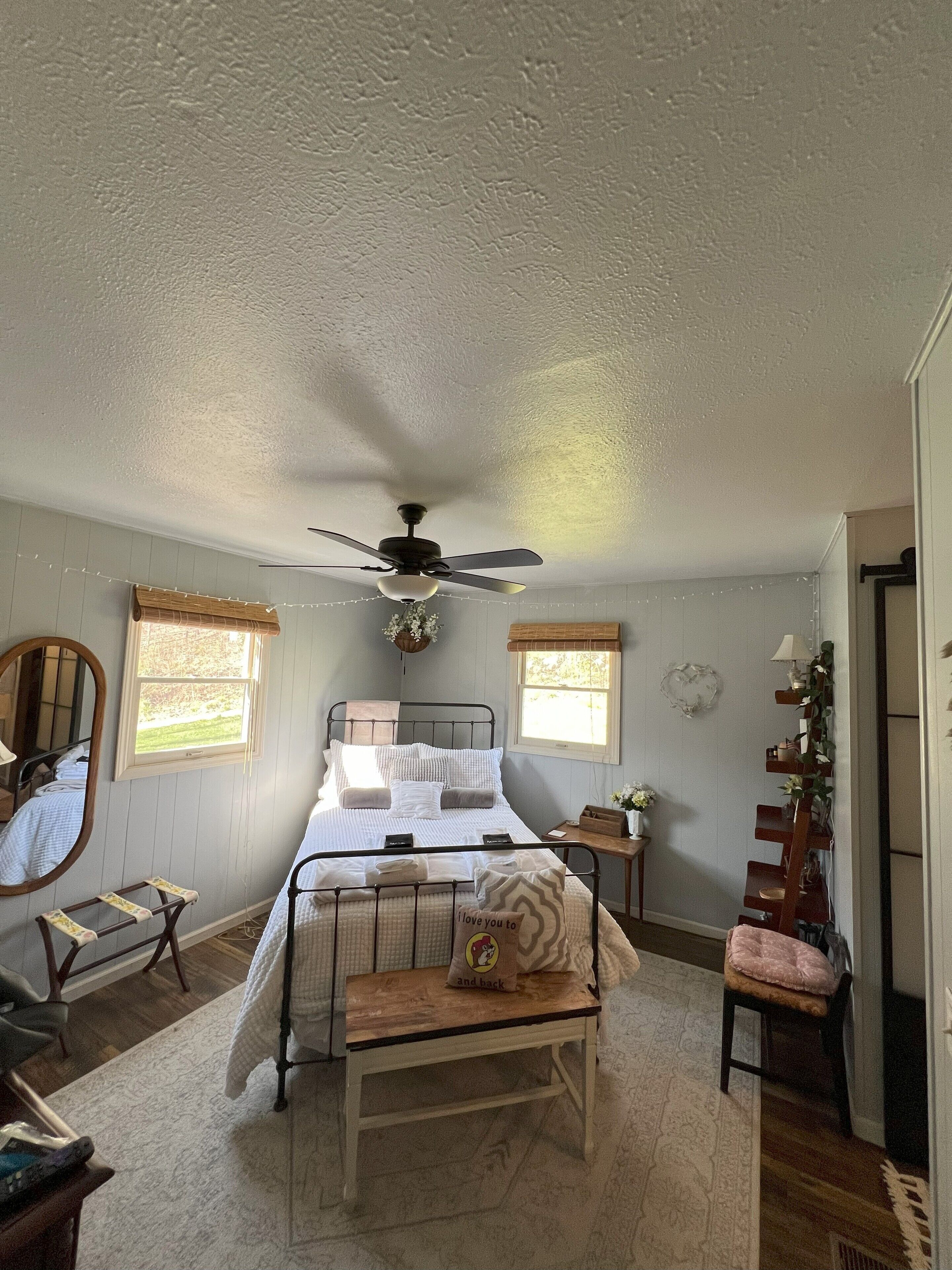 A bedroom with a bed, a ceiling fan, a bedside table, a chair, a mirror, and a window with blinds.