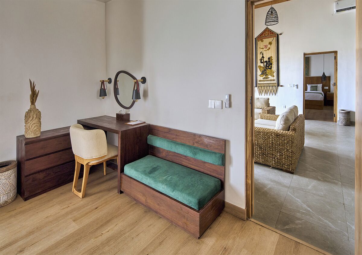 A room with a wooden desk, a green cushioned bench, a chair, a mirror, a decorative wall hanging, and a wicker sofa.