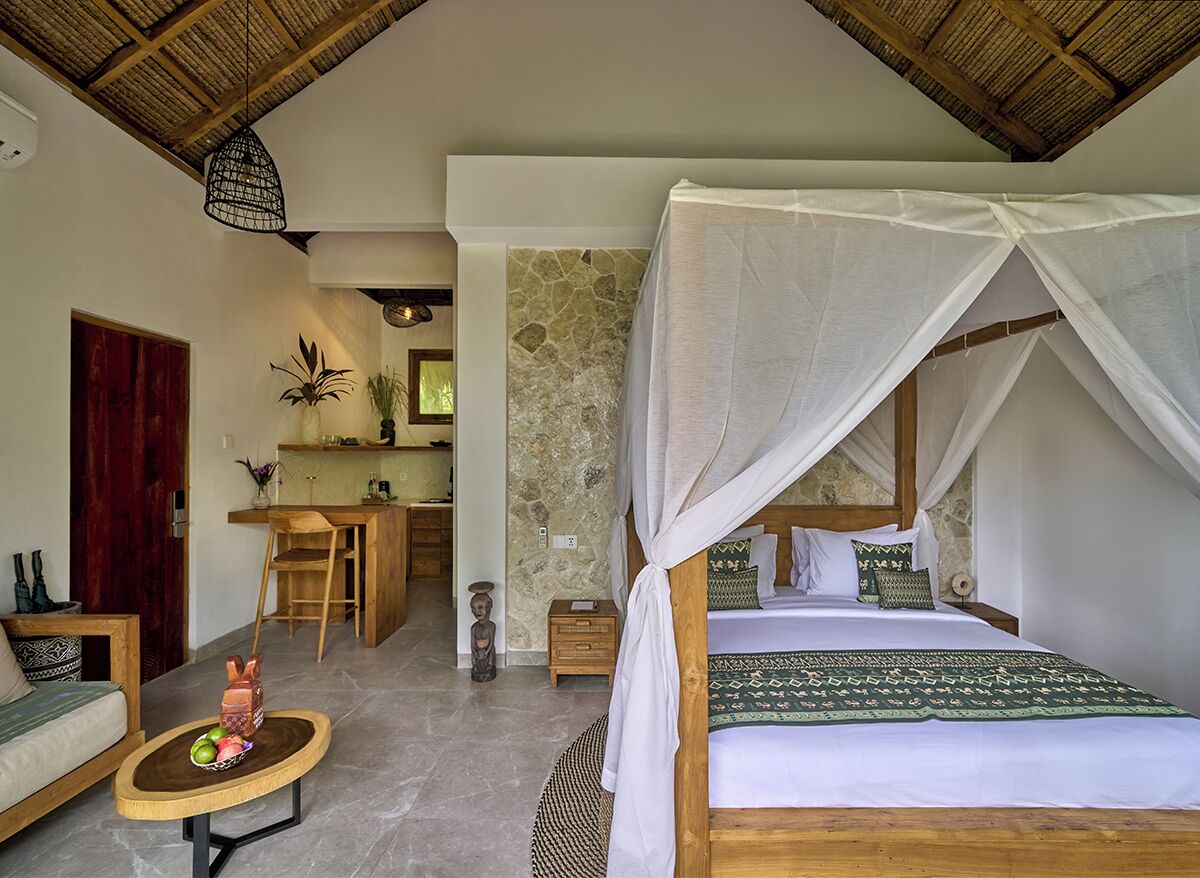 A bedroom with a four-poster bed, a sitting area with a table and chairs, and a kitchenette in the background.