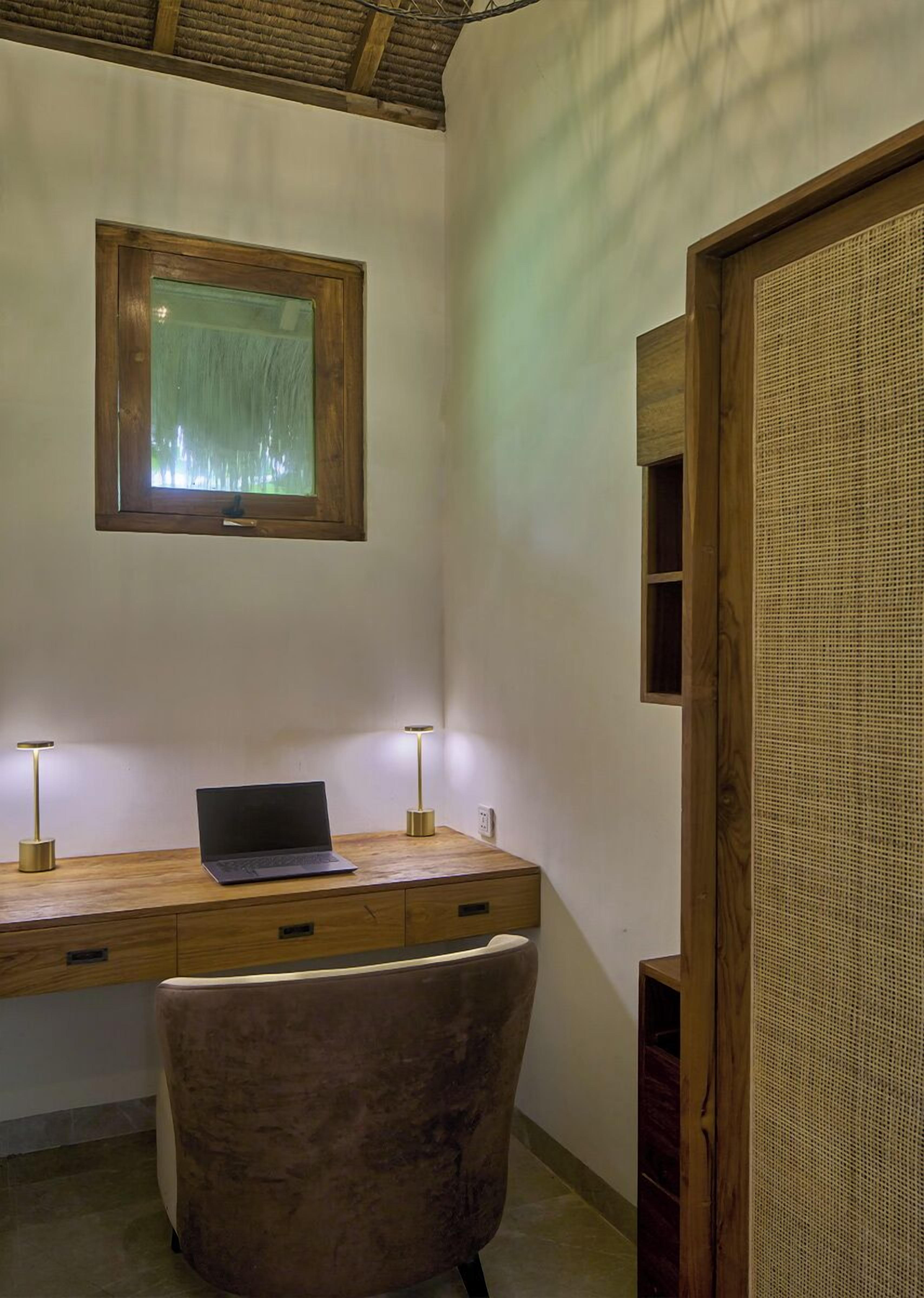 A wooden desk with a laptop, two lamps, a chair, and a window with a view.