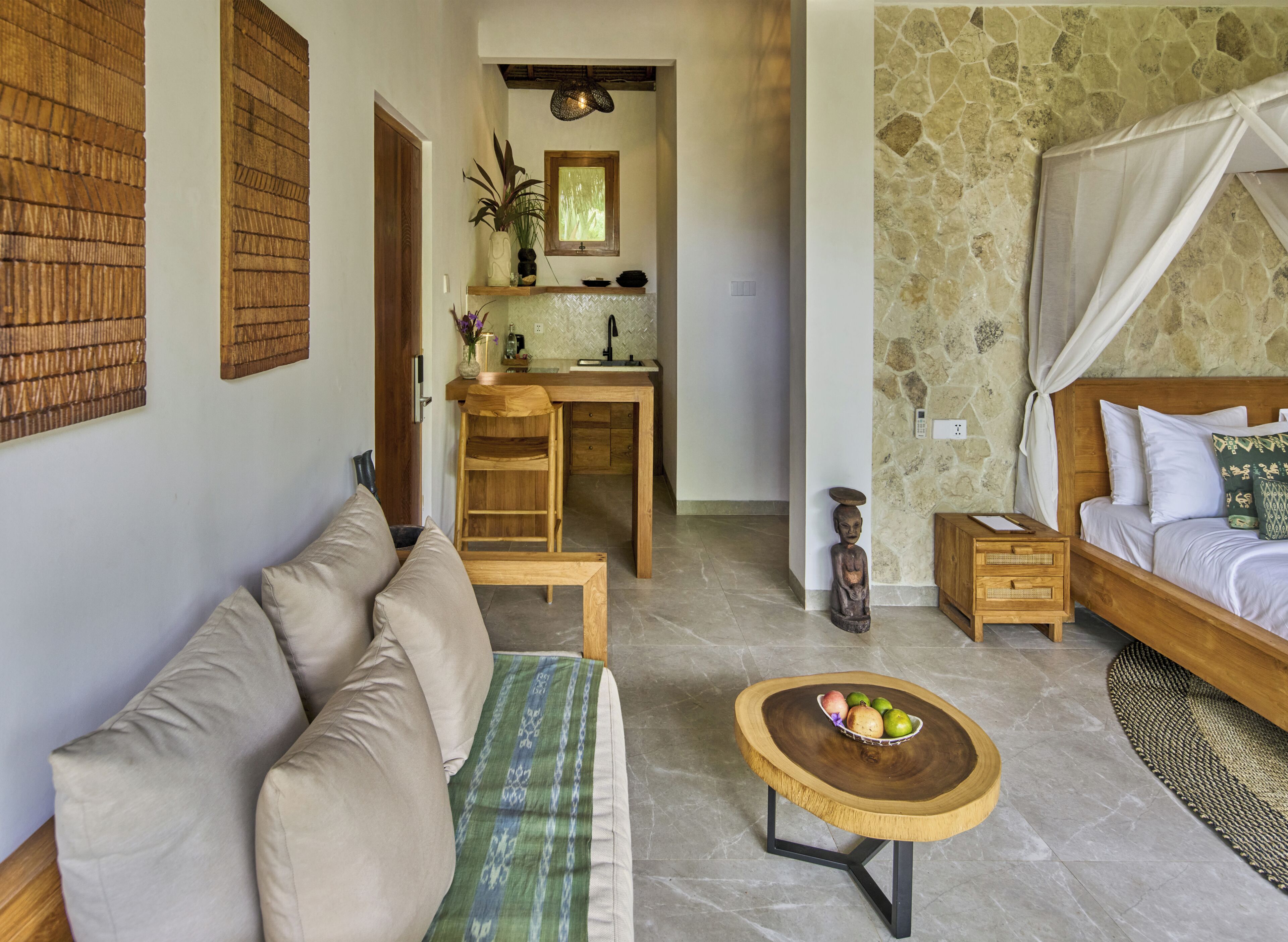 A hotel room with a sofa, a wooden table with fruit, a bed with a canopy, a stone wall, and a kitchenette.
