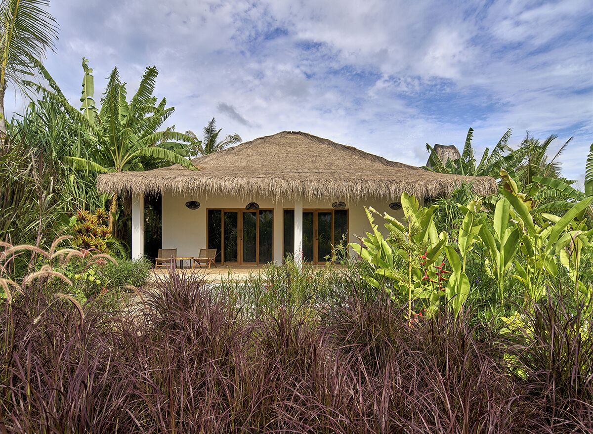 A bungalow with a thatched roof, surrounded by tropical vegetation.