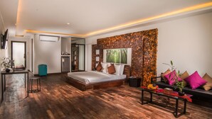 A modern hotel room with a wooden floor, a bed with pillows, a sofa with cushions, a small table, and a wall with a nature-themed artwork. - Elysium Suite (Siem Reap)