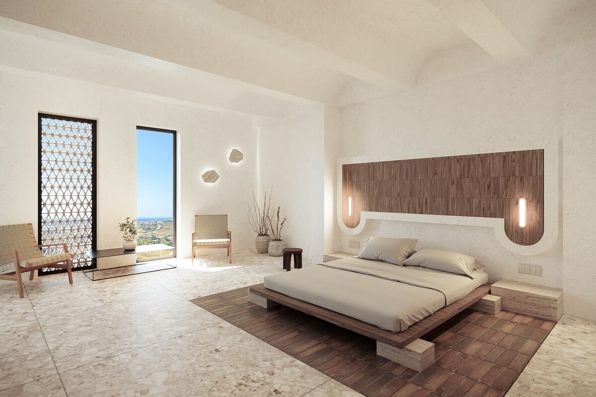A modern bedroom with a large bed, a wooden headboard, a seating area with a bench, and a view of the landscape through a large window.