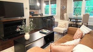 Una sala de estar moderna con un televisor de pantalla plana, una mesa de centro con un jarrón, un sofá con cojines y una zona de cocina con microondas y una mesa de comedor.