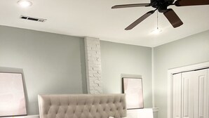 Un dormitorio con cabecera capitonada, un edredón blanco grande, dos mesitas de noche con lámparas y un ventilador de techo.