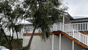 Une terrasse en bois avec une balustrade blanche, un sol sablonneux et une clôture blanche ornée d’un motif en treillis.