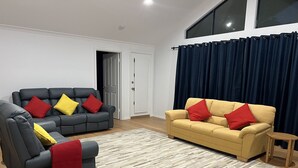 Un salon avec deux canapés, l’un gris et l’autre jaune, tous deux agrémentés de coussins rouges. Le sol est en bois et une rideau bleu foncé recouvre la fenêtre.