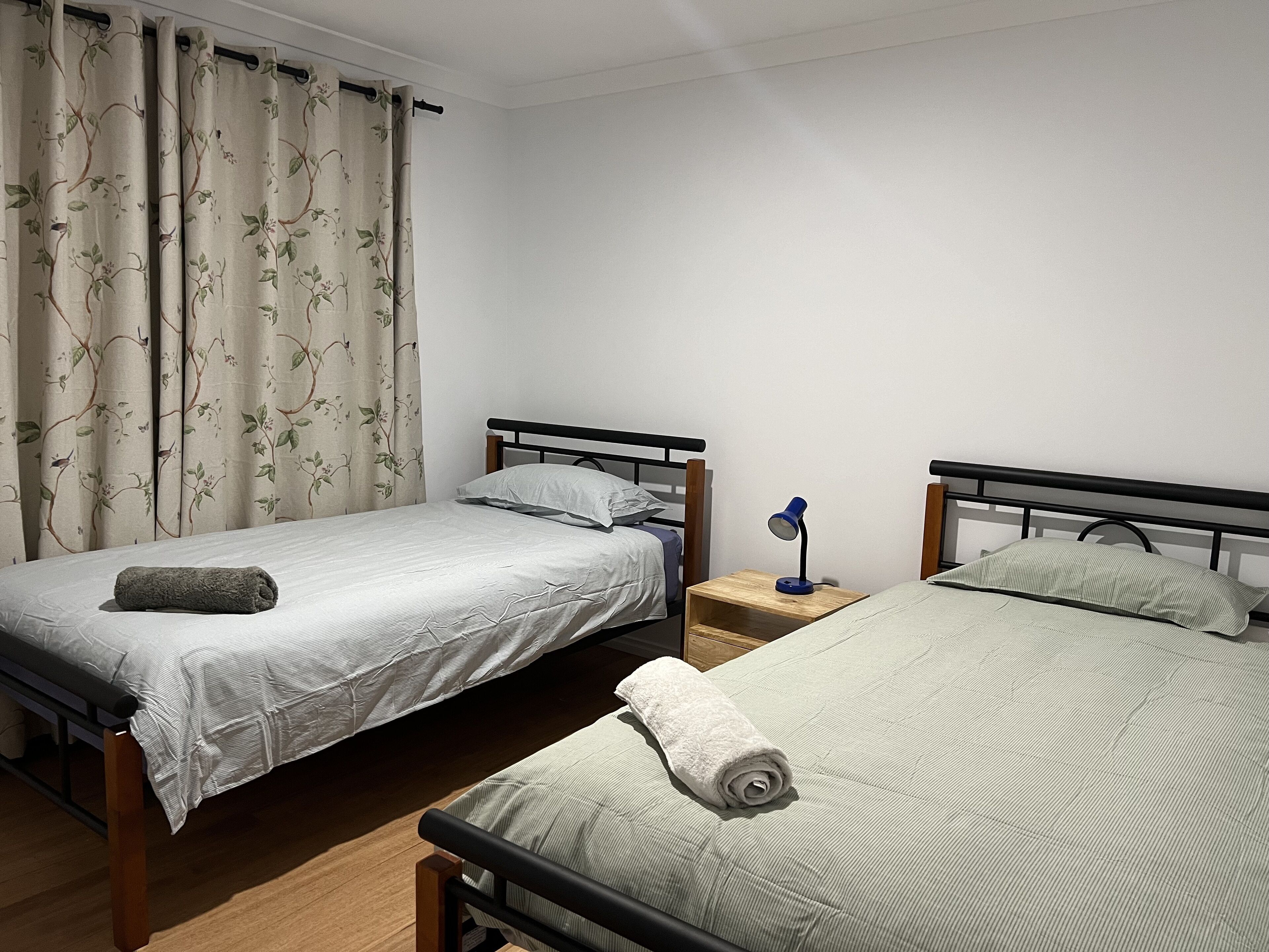 Una habitación con dos camas individuales, una mesita de noche con lámpara y una ventana con cortinas florales.