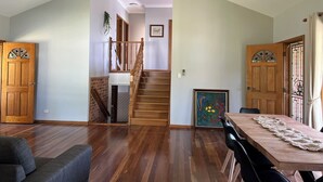 A spacious living area with a wooden staircase leading to an upper floor, a dining table with chairs, and a large framed artwork on the wall.