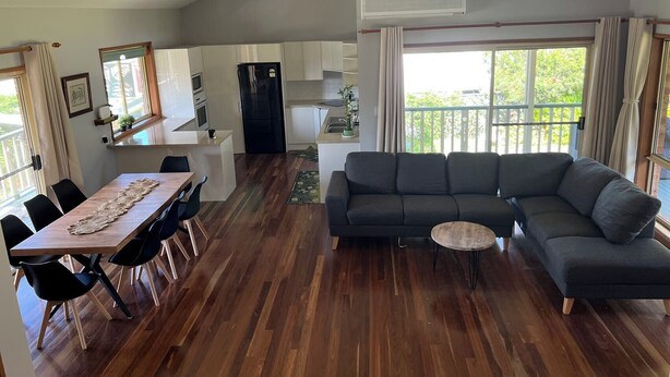 A spacious living area with a dark gray sectional sofa, a wooden dining table with chairs, a small round coffee table, and a kitchen in the background.
