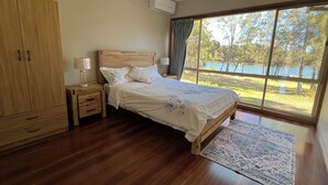 A bedroom with a wooden bed, a nightstand, a wardrobe, and a large window with a view of a lake.