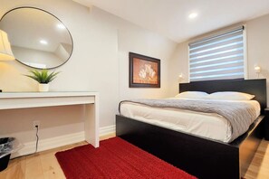 A bedroom with a bed, a bedside table, a mirror, a framed picture, and a window with blinds.