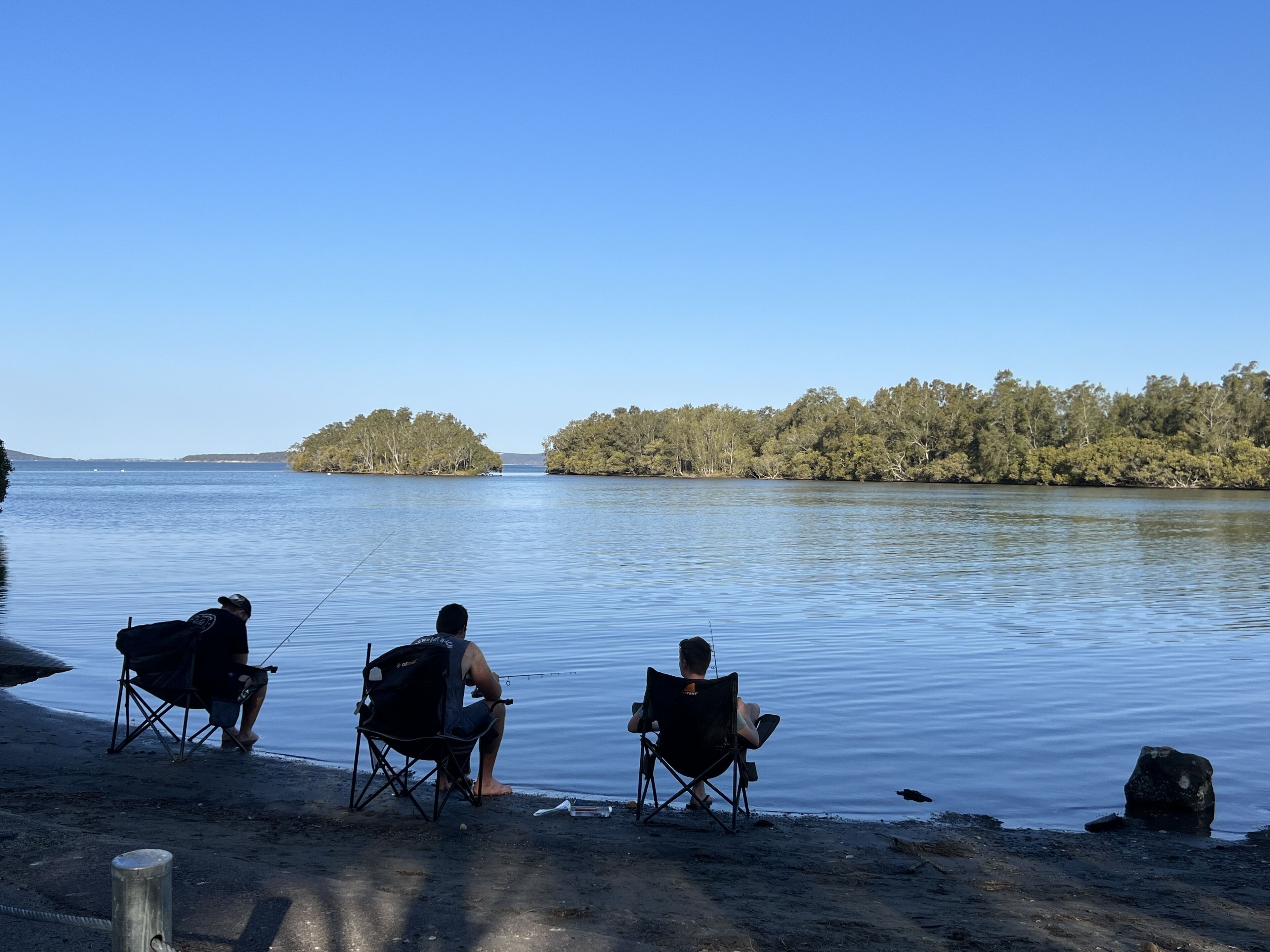 Two people fishing by a calm lake with a forested island in the background.