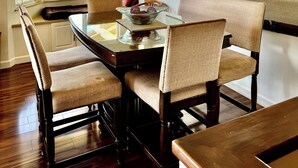 A dining area with a wooden table and four upholstered chairs, a vase with flowers, and a view of the outdoors through large windows.