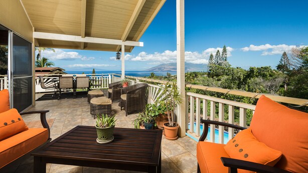A covered outdoor seating area with wicker furniture, potted plants, and a view of the ocean and mountains.