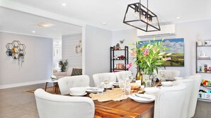 Un coin repas avec une table en bois dressée pour six personnes, entourée de chaises blanches. On y trouve une décoration murale hexagonale, une bibliothèque remplie de divers objets et un tableau représentant un paysage accroché au mur.