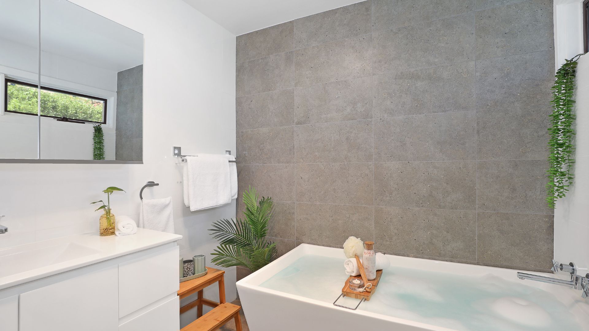 Een moderne badkamer met een ruime badkuip, een witte wastafel, een spiegel en een grijze betegelde muur.
