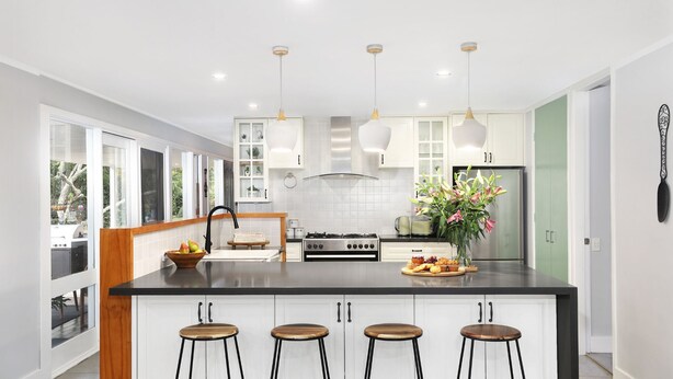 Une cuisine moderne avec des meubles blancs, un îlot central doté de tabourets de bar, une cuisinière en acier inoxydable et des luminaires suspendus.