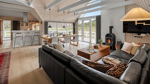 Amplia sala de estar con un sofá de cuero negro grande, una mesa de centro de madera y una chimenea. Hay un comedor con mesa y sillas, y una cocina con alacenas blancas y un ventanal.
