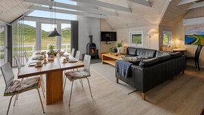 Amplia sala de estar con techo de madera, un sofá negro, una mesa de comedor preparada para cuatro personas y una chimenea.