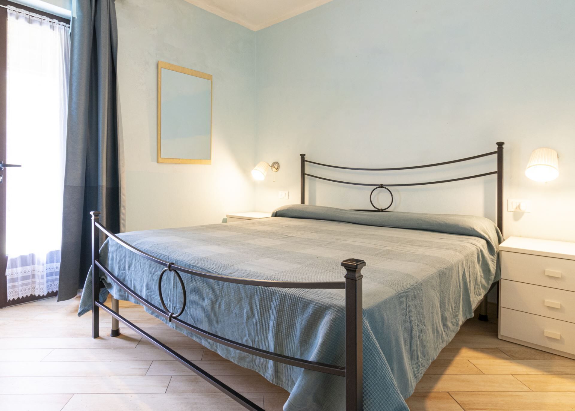 Quarto com estrutura de cama em metal, mesa de cabeceira, luminária de parede e criado-mudo.