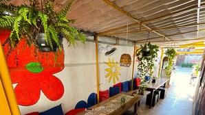 Un espace extérieur abrité, aménagé avec une longue table en bois, des bancs dotés de coussins colorés, des plantes suspendues et un sol carrelé.