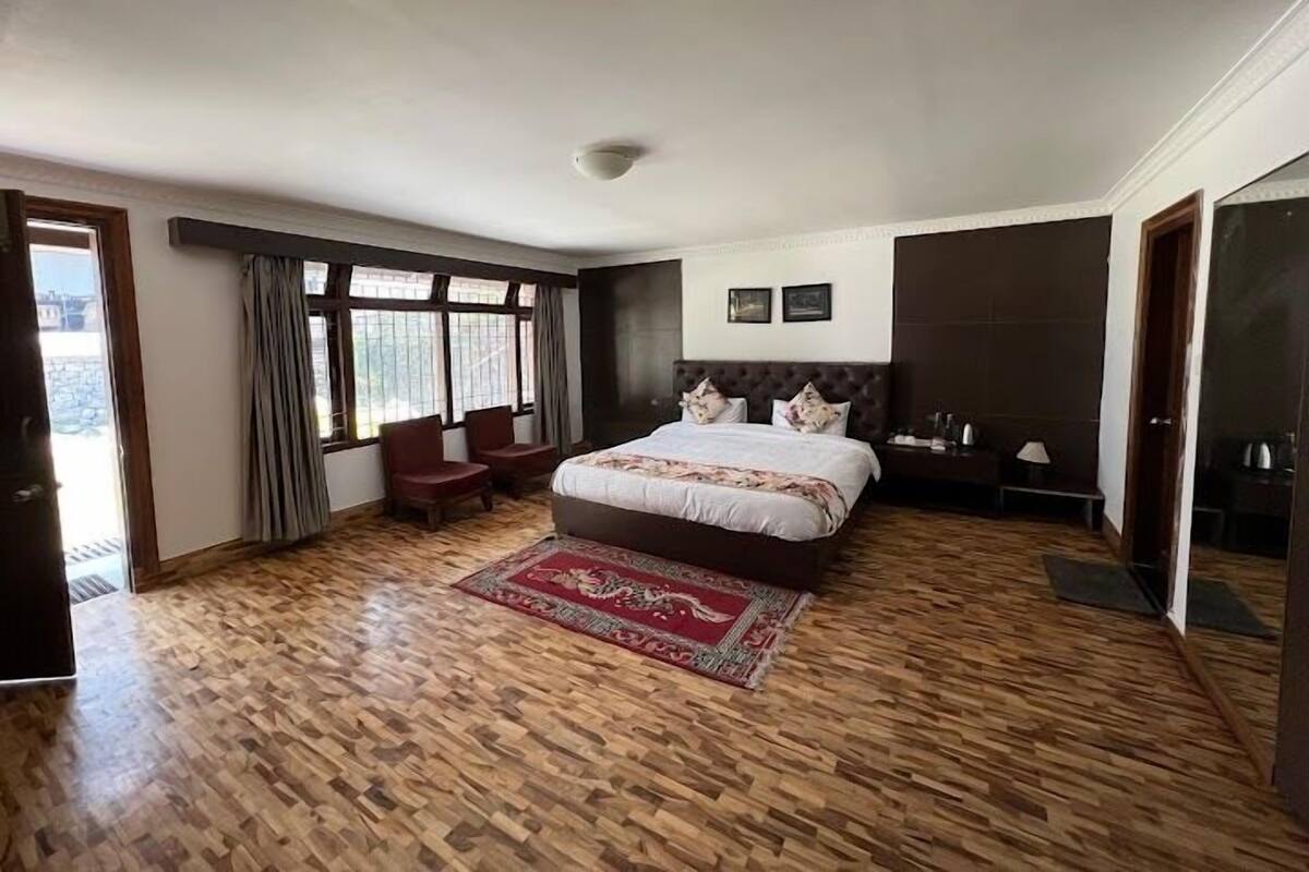 A spacious bedroom with a large bed, a wooden floor, a red rug, a seating area with chairs, and a view through the window.