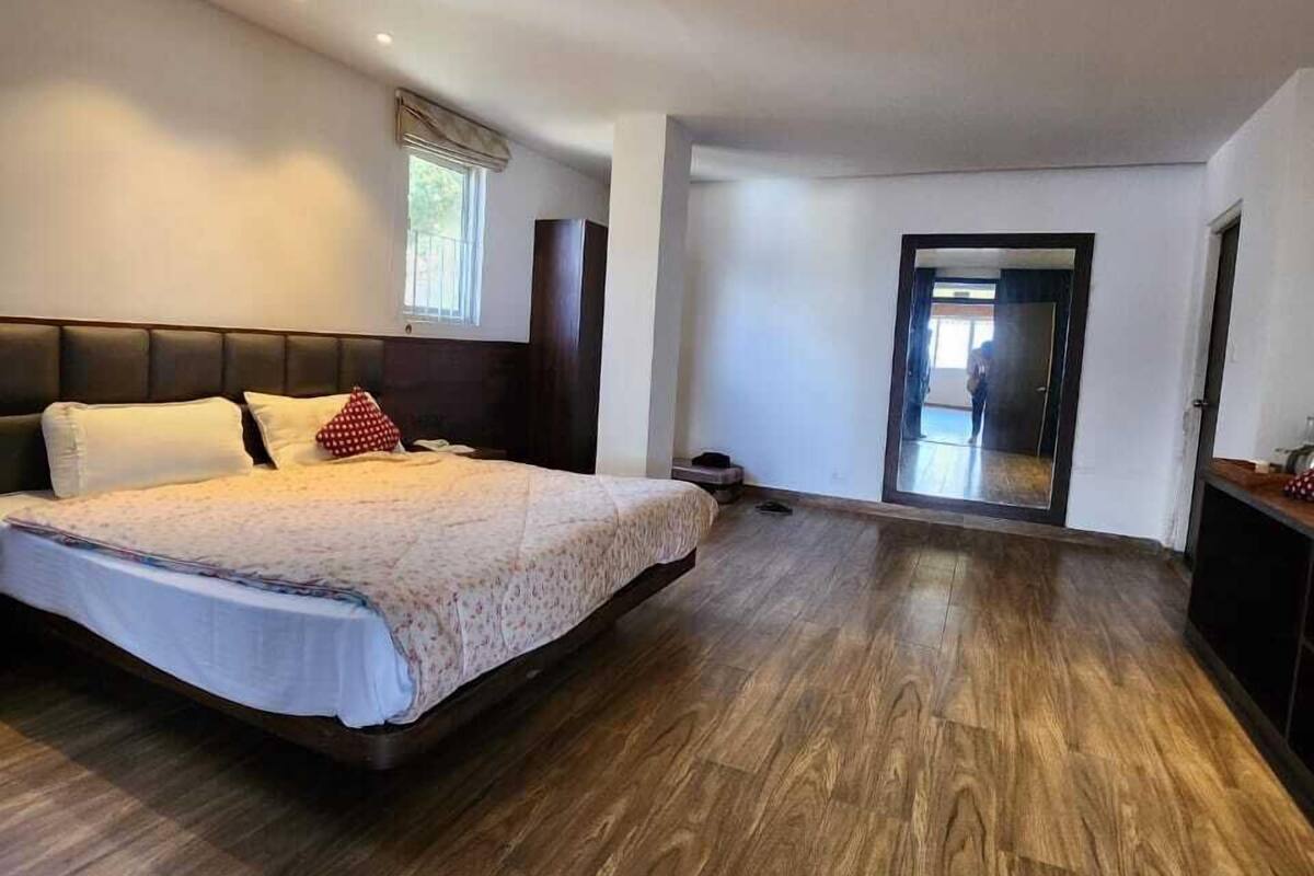 A bedroom with a large bed, wooden flooring, a window with blinds, and a door leading to another room.