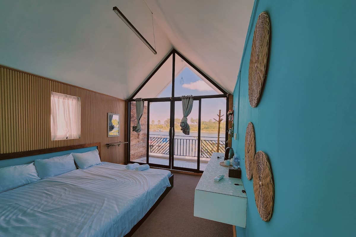 A bedroom with a large bed, a small table with a lamp, a window with curtains, and a view of the outdoors.