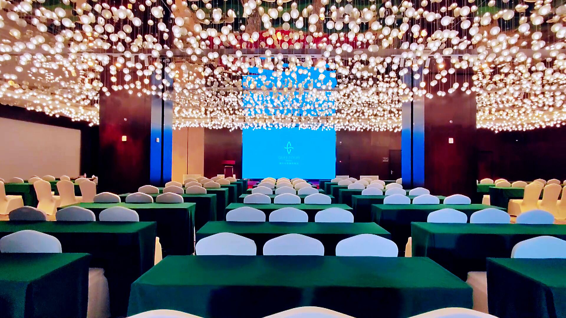 A conference room with rows of tables and chairs, a large screen at the front, and decorative hanging lights.