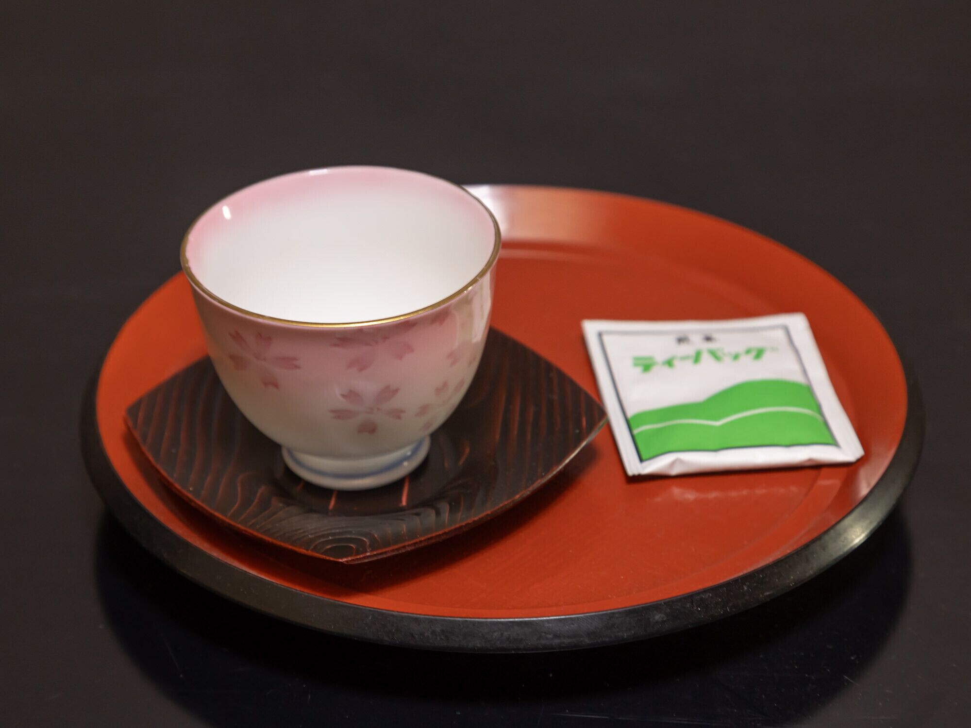 A teapot with a floral pattern on a saucer, placed on a red tray with a packet of tea beside it.