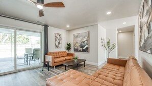 En moderne stue med en stor sofa, en mindre sofa, et sofabord og et spiseområde med bord og stole. Der er en ventilator i loftet, et billede på væggen og en plante.