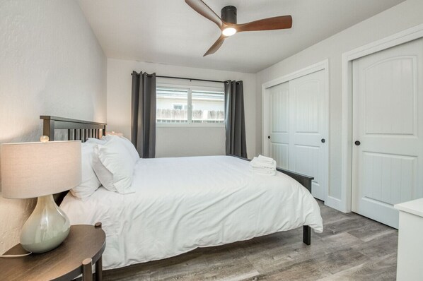 Une chambre à coucher avec un lit, un ventilateur de plafond, une table de chevet, une lampe et une fenêtre avec des rideaux.
