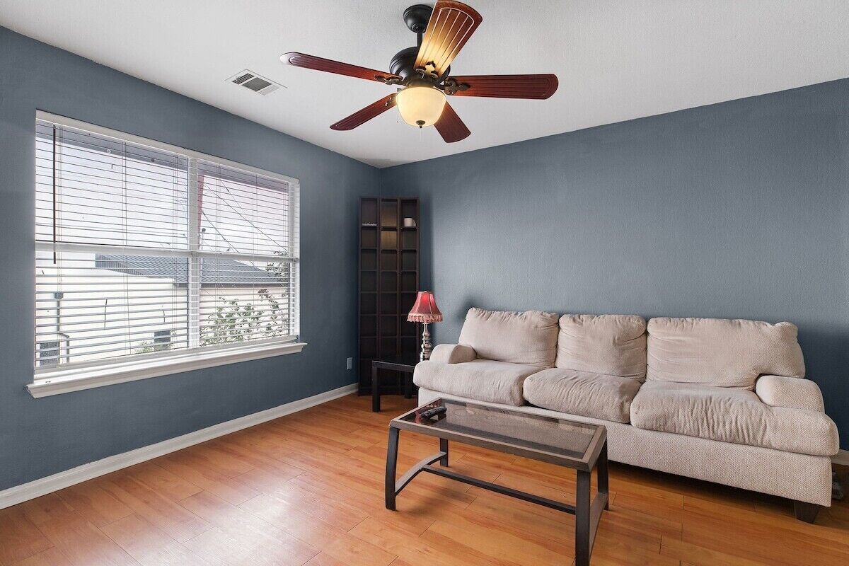 A living room with a beige sofa, a glass coffee table, a ceiling fan, and a window with blinds.