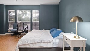 Une chambre à coucher avec un lit, un bureau, une chaise et une fenêtre avec des stores.