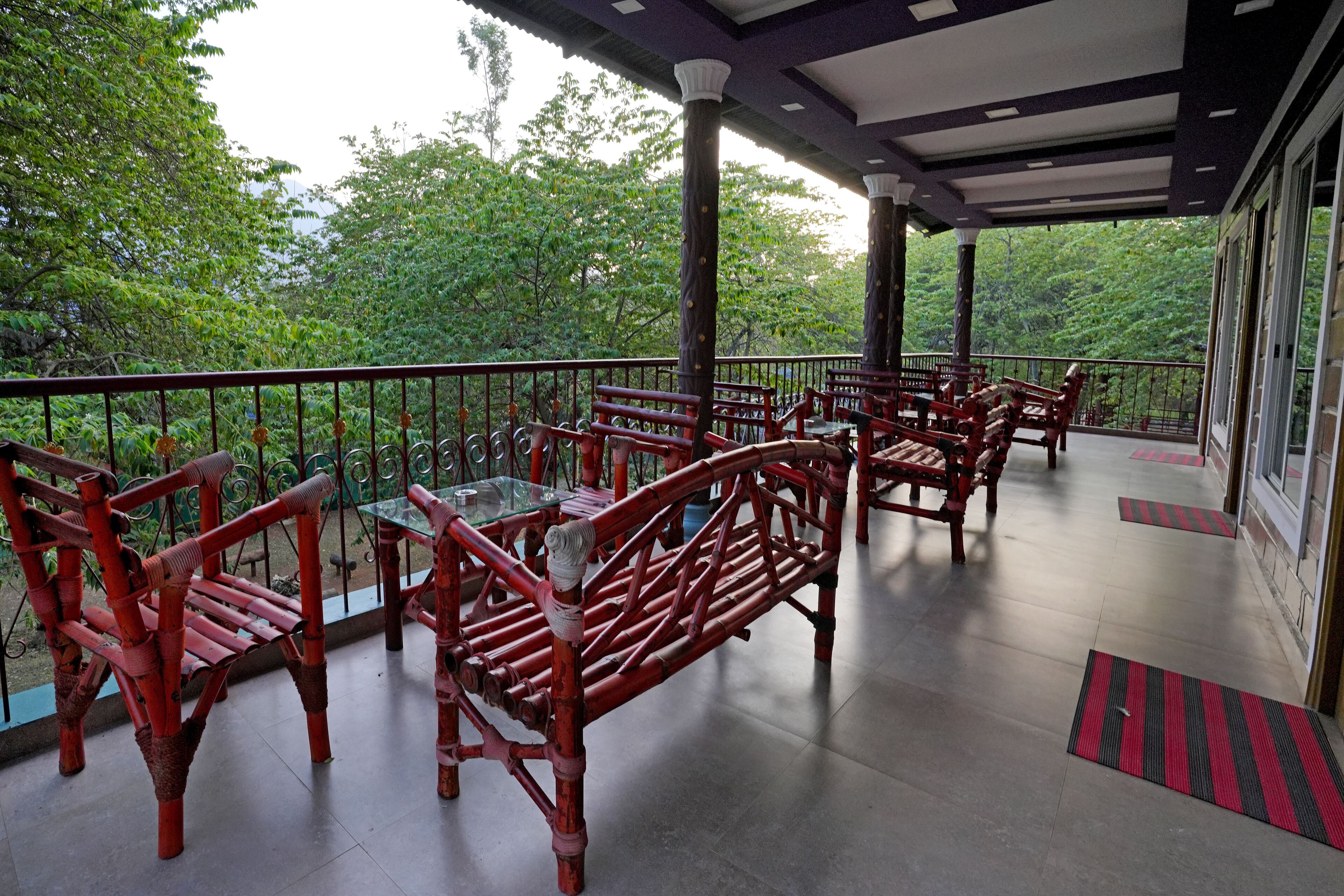 A covered outdoor seating area with red wooden furniture and glass tables, surrounded by greenery.