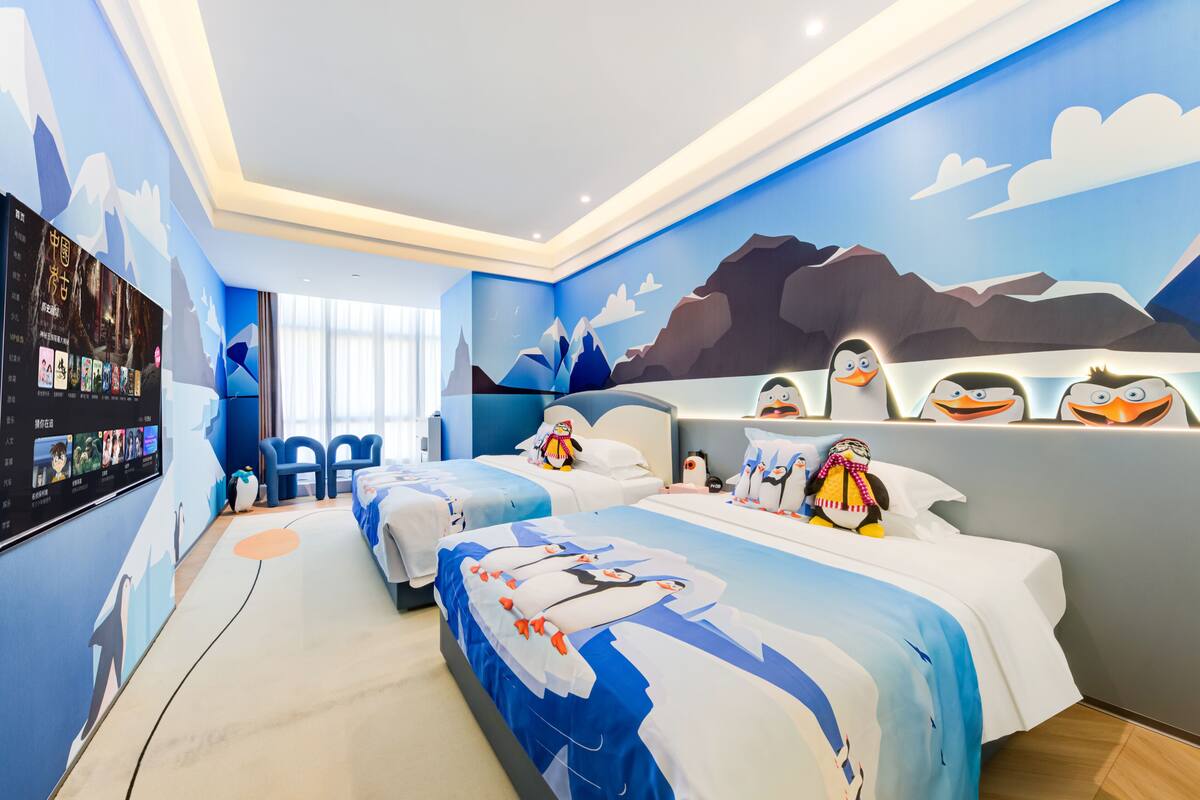 Quarto de hotel com um grande mural de montanhas e pinguins na parede, duas camas com roupa de cama combinando, uma televisão de tela plana na parede à esquerda e um pequeno espaço de estar com cadeiras.