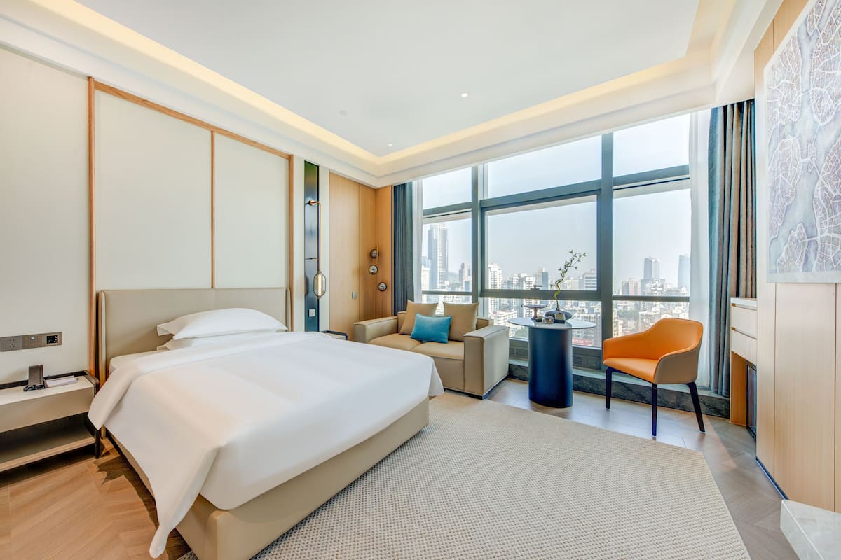 Quarto de hotel moderno com uma cama grande, um sofá, uma cadeira e vista para a cidade através de grandes janelas.