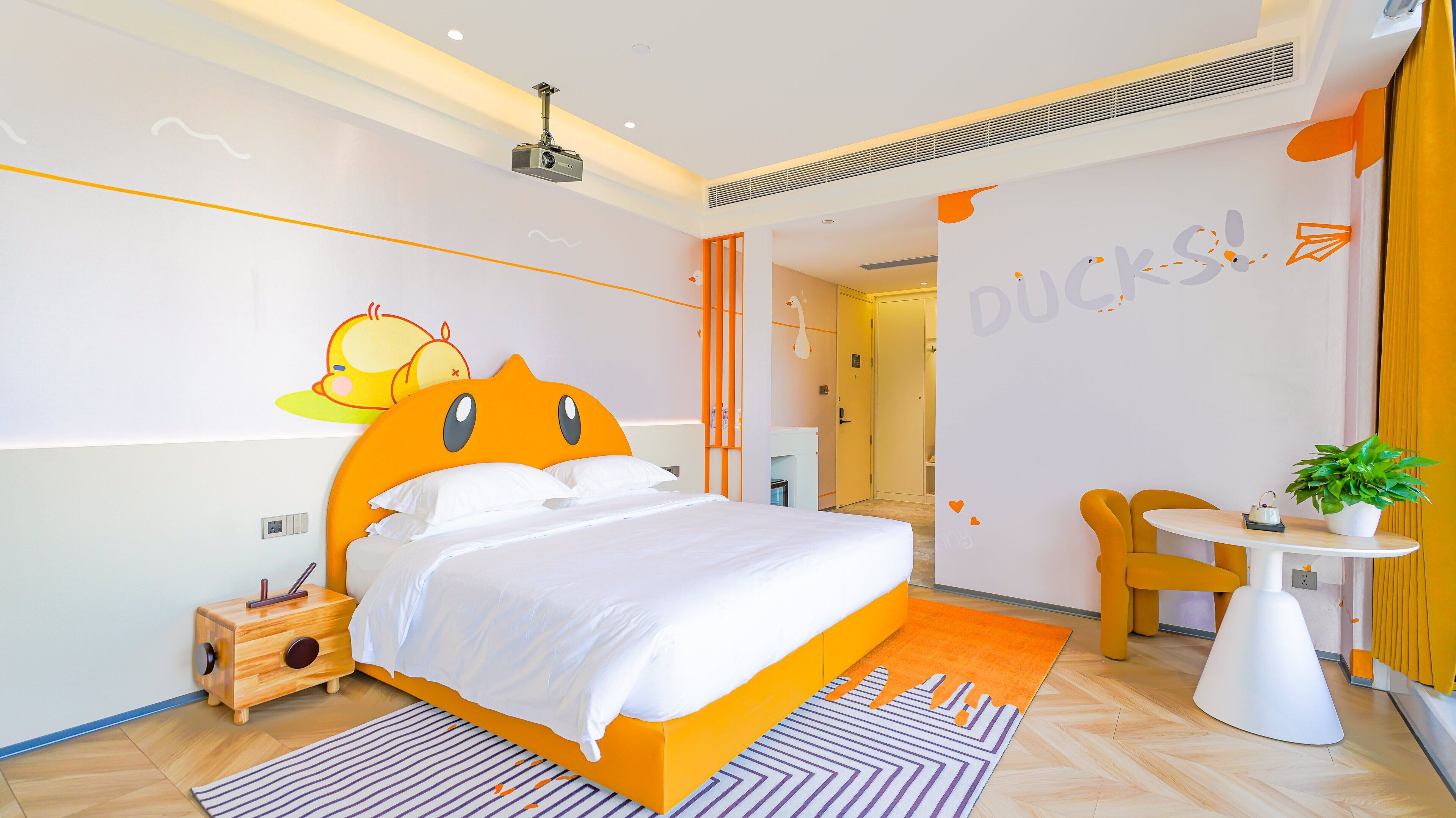 A modern bedroom with a large bed, a small bedside table, a chair, a table with a plant, and a projector on the ceiling.