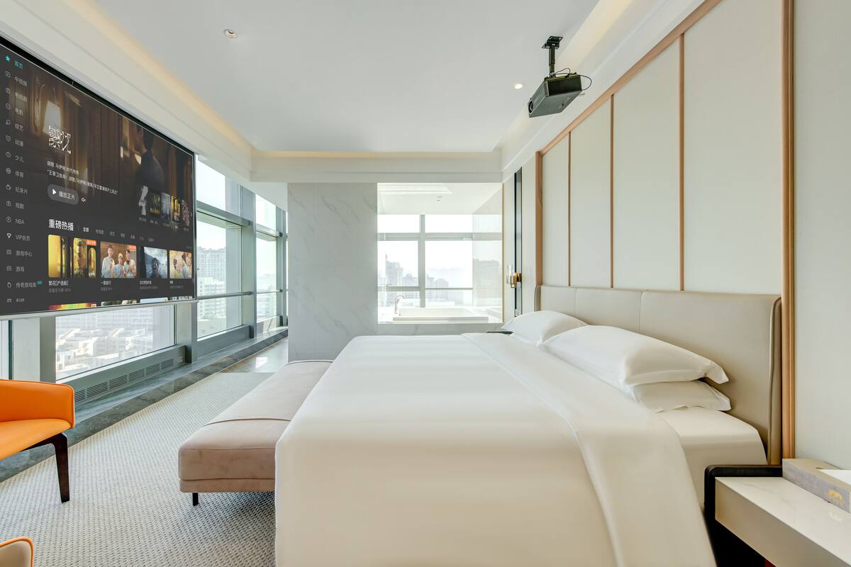 Quarto de hotel moderno com uma cama grande, área de estar e uma tela exibindo conteúdo.