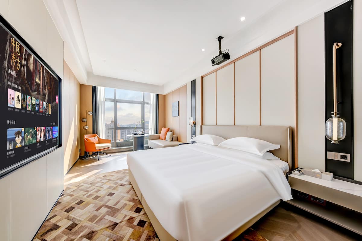 Quarto de hotel moderno com uma cama grande, televisão de tela plana, área de estar com sofá e poltrona, e vista para a cidade por grandes janelas.