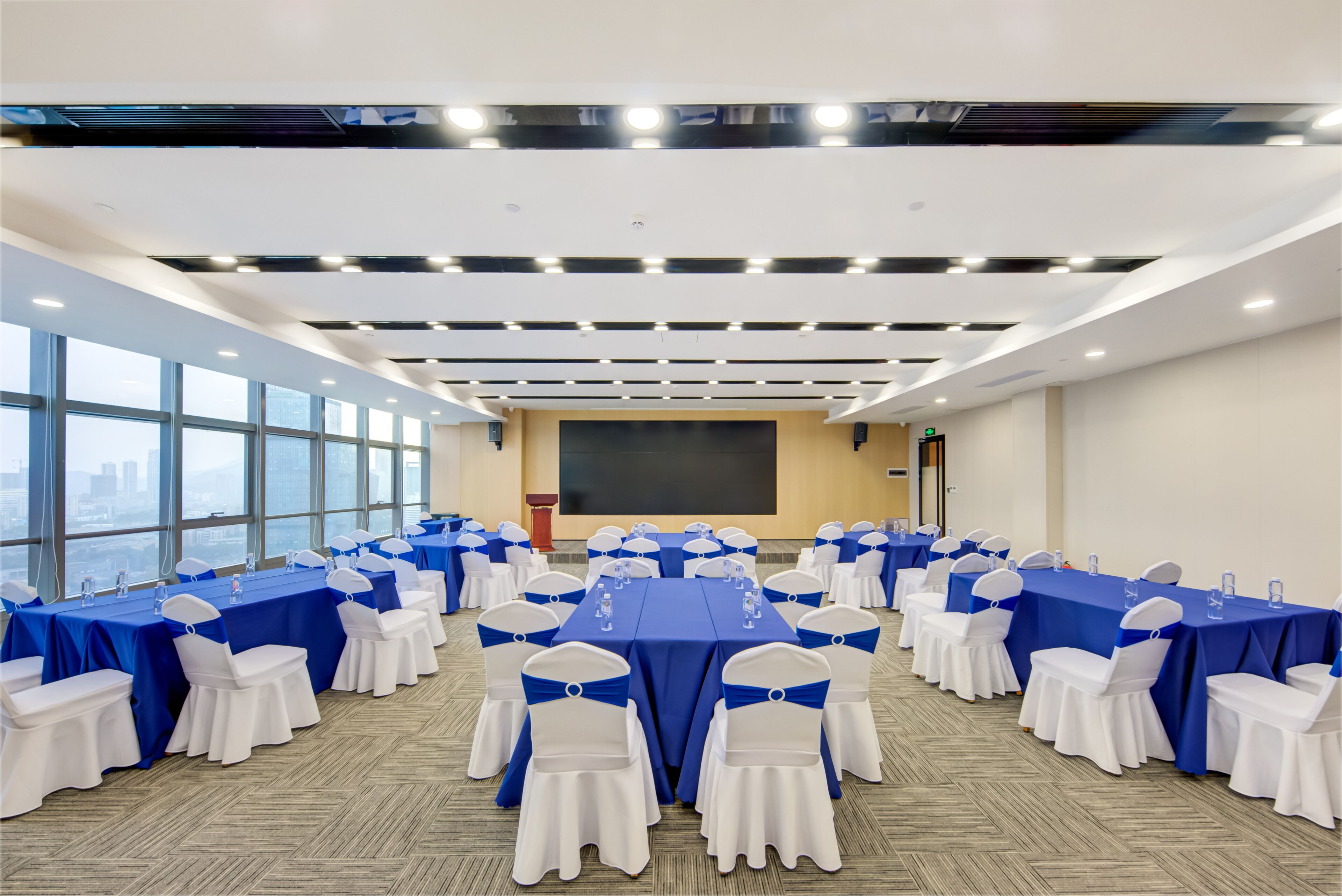 Une salle de conférence dotée de plusieurs tables et chaises, de grandes fenêtres et d’un écran de projection.