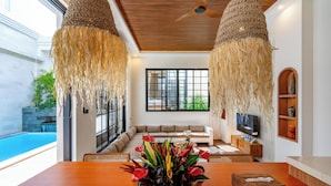 Un salon moderne avec un plafond en bois, une grande fenêtre, un canapé, un téléviseur et une table à manger agrémentée d’un centre de table floral.