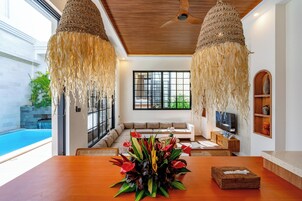 Una sala moderna con techo de madera, un ventanal grande, un sofá, un televisor y una mesa de comedor con un centro de mesa floral.